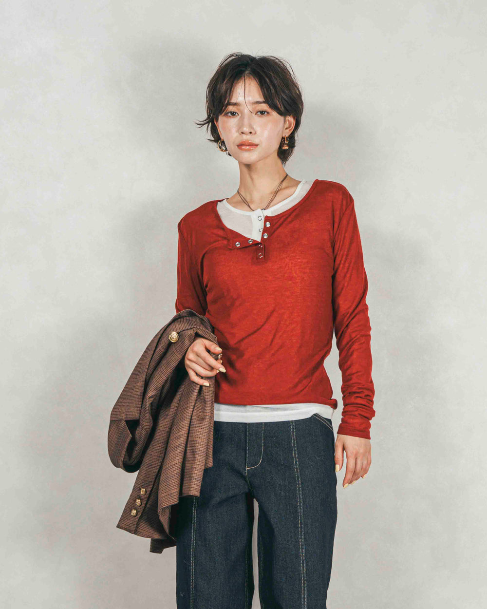 白のシアーシャツの上に赤のヘンリーネックトップスを重ね着し、デニムパンツとブラウンのジャケットを合わせた、おしゃれなレイヤードコーディネート。｜KaLae